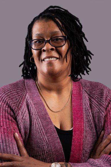Smiling woman with glasses, wearing a pink cardigan, crossed arms against a gray background.